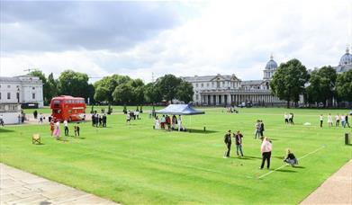 Red bus and people stood on green grass on the Royal Museums Greenwich Grounds
