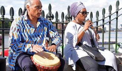 Photo of a man in a blue and white printed shirt sitting playing a drum next to a woman sitting beside him singing into a microphone wearing a lilac h