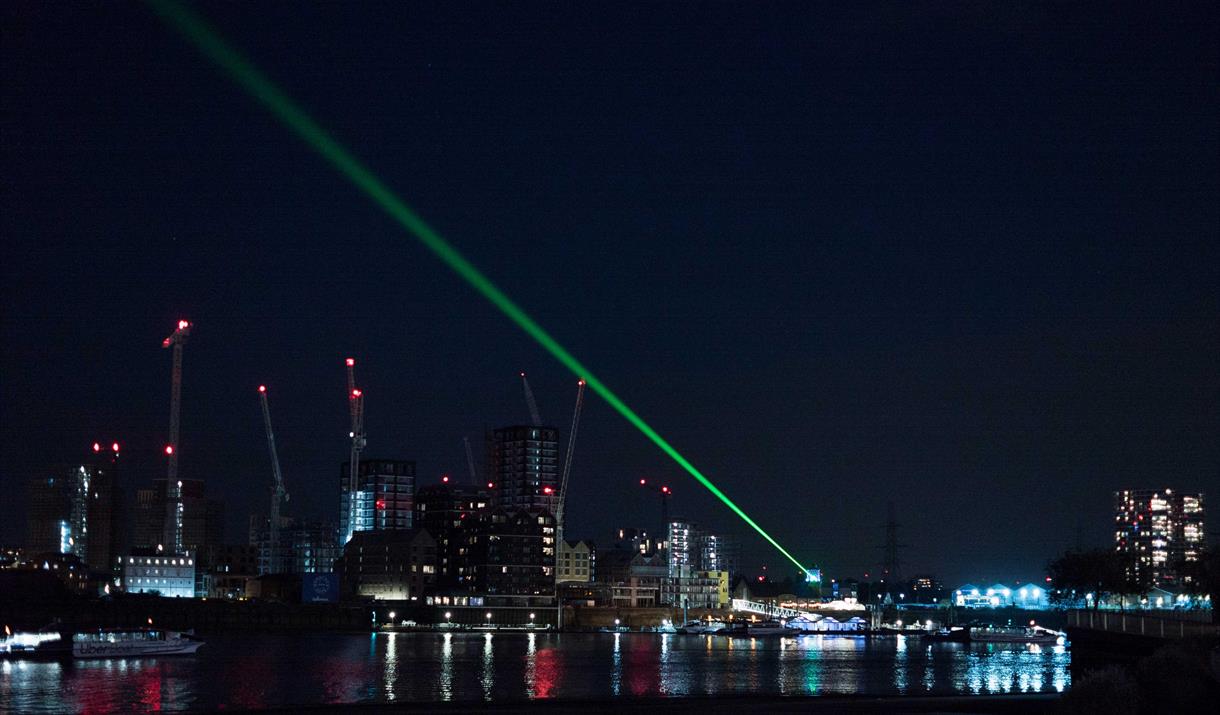 A view of the Thames river in London at night, with tower blocks and cranes shown on the far bank.  From a lighthouse in the distance a green laser sh