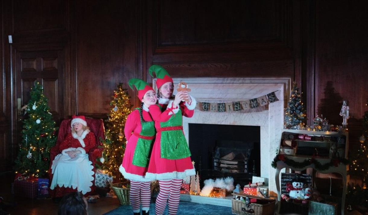 Two actors dressed as elves are performing in a cozy Christmas room.