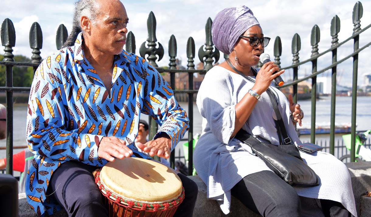 Photo of a man in a blue and white printed shirt sitting playing a drum next to a woman sitting beside him singing into a microphone wearing a lilac h