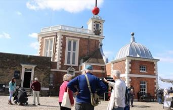 Royal Observatory made of red bricks and white windows and railing, with red timeball on the roof and Tour Guide and visitors around.