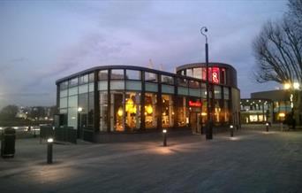 Outside Nando's next to The Cutty Sark in Greenwich, showing a modern building with lots of seating and a view onlooking the river Thames.