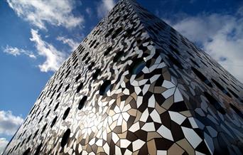 Exterior image of Ravensbourne University London with the building swathed in different coloured, tesellated aluminium tiles and circular windows of d