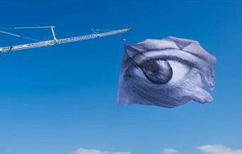 Dan Acher’s We Are Watching is a striking image of a giant eye flown on a monumental flag, comprising of thousands of digital portraits.