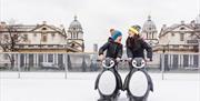 Two children laughing as they hold onto model penguins to steady themselves on the ice at the Queen's House Ice Rink in Greenwich.