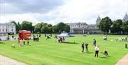 Red bus and people stood on green grass on the Royal Museums Greenwich Grounds
