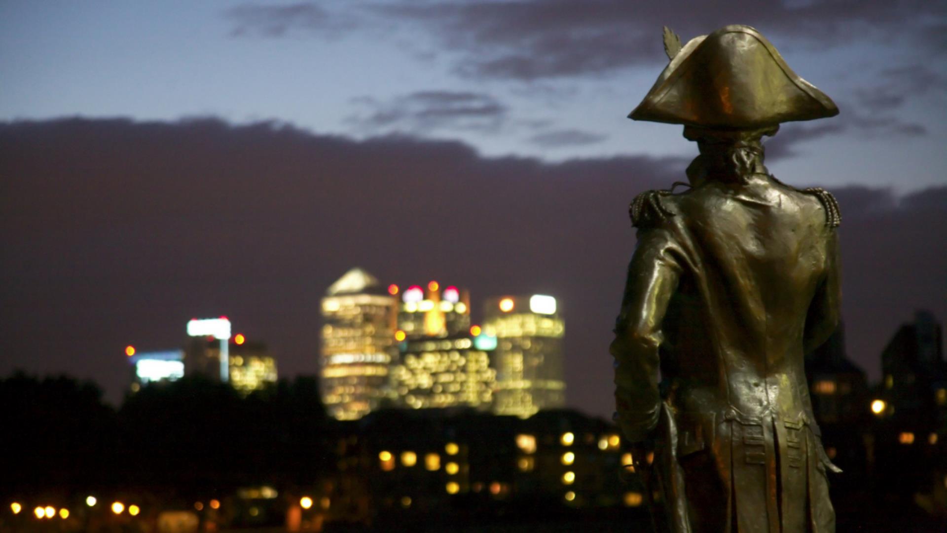 A statue of Lord Nelson overlooks London Docklands at night.