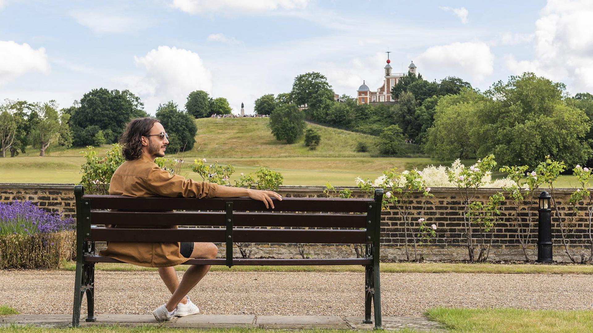 A man sits on a bench overlooking Greenwich Park and the Royal Observatory.