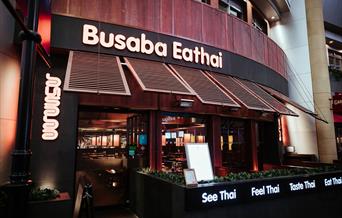 Busaba Eathai entrance in wood finish with wooden windows which are open and has the restaurant name in beige on top. Also has outside seating.