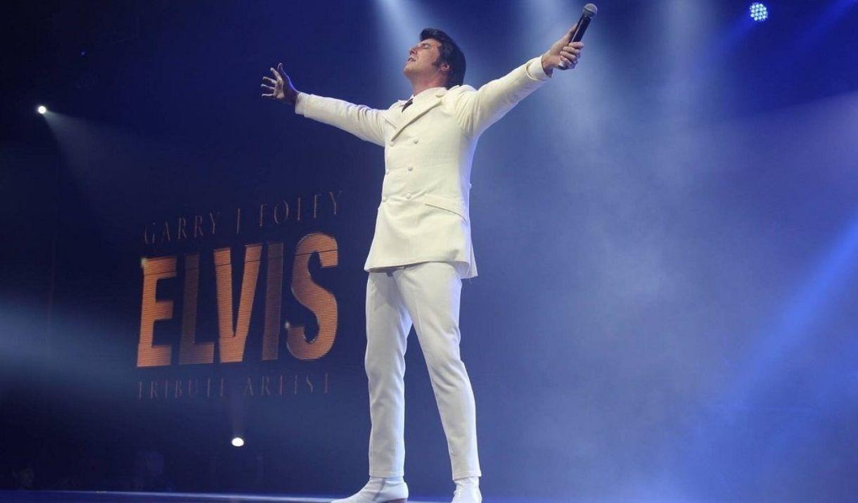 Elvis lookalike wearing white outfit performing onboard City Cruises Elvis Tribute Cruise with blue background.
