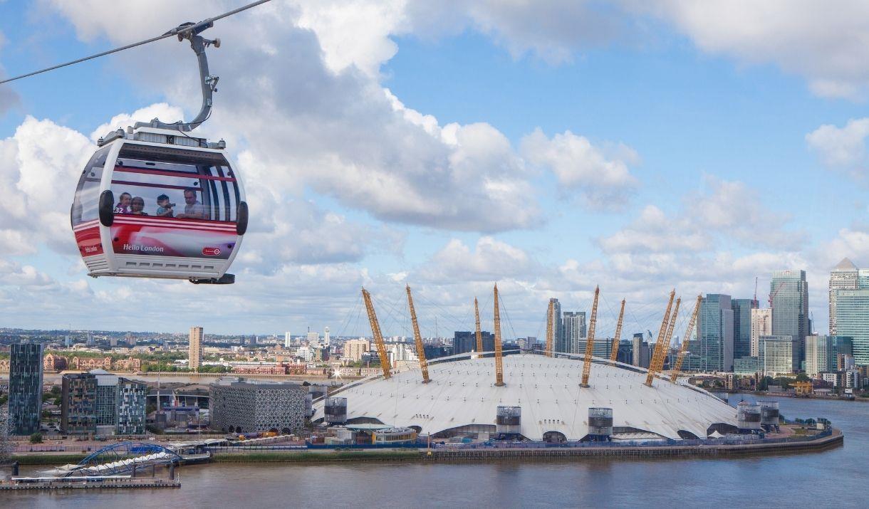 Emirates Air Line carrier on top of river Thames, overlooking The O2 which is a white dome with 12 yellow pillars around on top.