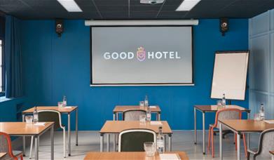 Good Hotel, the blue room. A room with blue walls, grey flooring, multiple single person desks with single chairs, a projector board and a business bo