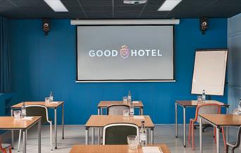Good Hotel, the blue room. A room with blue walls, grey flooring, multiple single person desks with single chairs, a projector board and a business bo