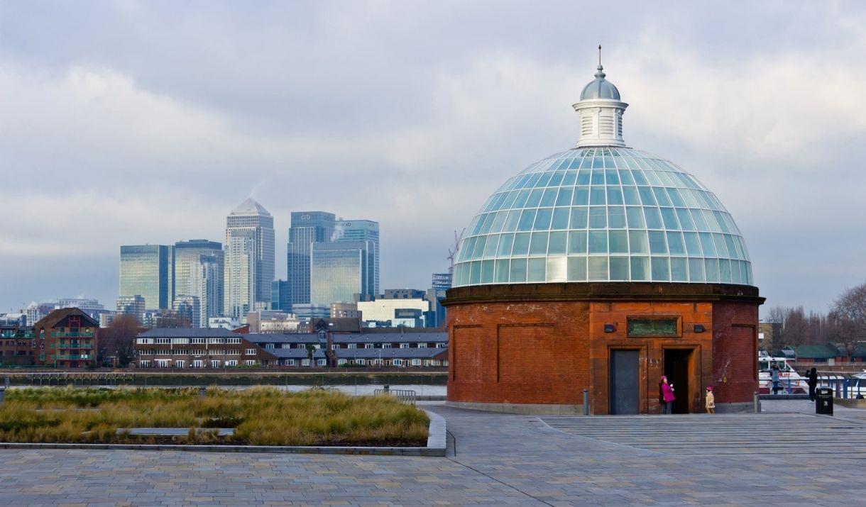 Image shows Greenwich Foot Tunnel made of red bricks with a dome made of matte finished glass.  In the background you can Canary Wharf skyline.