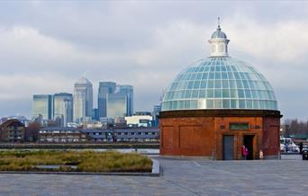 Image shows Greenwich Foot Tunnel made of red bricks with a dome made of matte finished glass.  In the background you can Canary Wharf skyline.