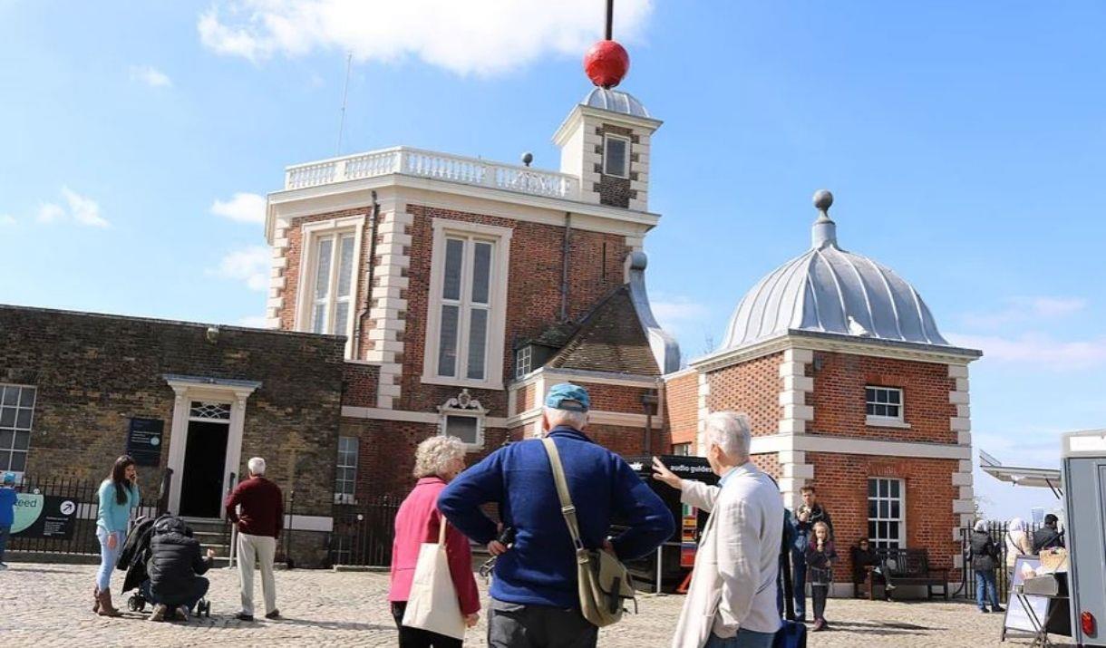 Royal Observatory made of red bricks and white windows and railing, with red timeball on the roof and Tour Guide and visitors around.