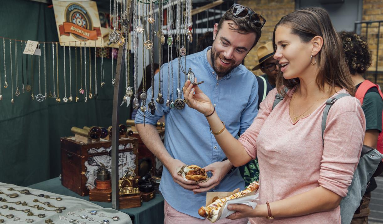 A couple admire the jewellery on a stand at Greenwich Market with some fresh street food in-hand.