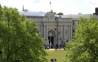 The grand exterior of the National Maritime Museum in Greenwich, surrounded by beautiful trees and a family having a picnic outside.