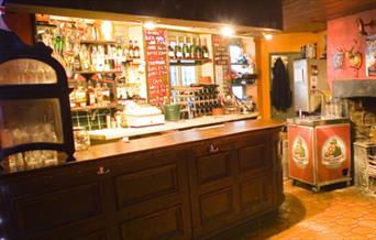 A photo of the bar area at Olivers Jazz Bar, The Bar is wooden and behind it is shelfs filled with bottles. To the right of the bar is a small old fa