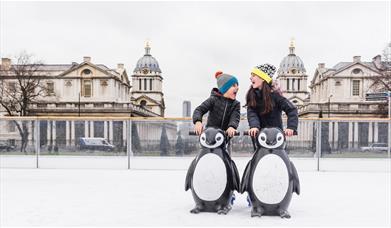 Two children laughing as they hold onto model penguins to steady themselves on the ice at the Queen's House Ice Rink in Greenwich.