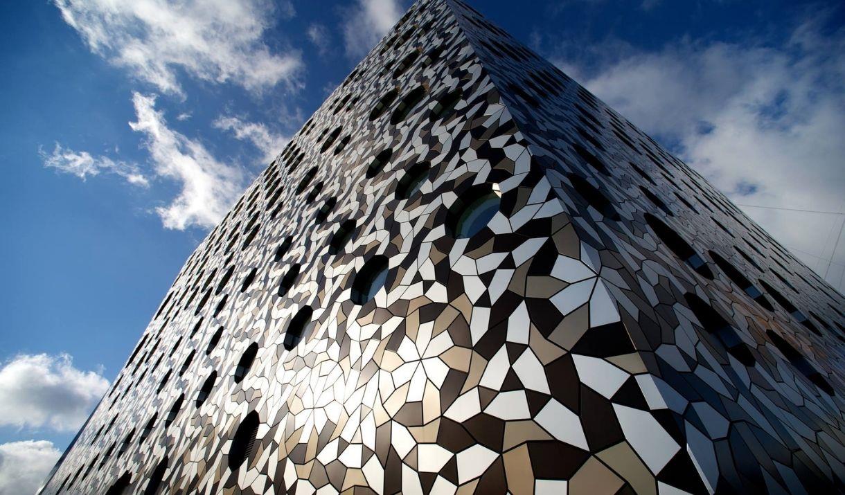 Exterior image of Ravensbourne University London with the building swathed in different coloured, tesellated aluminium tiles and circular windows of d