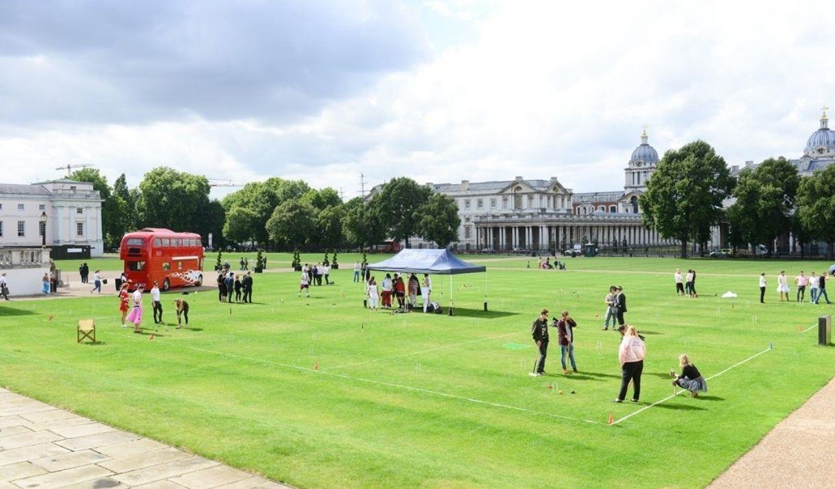Red bus and people stood on green grass on the Royal Museums Greenwich Grounds
