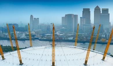 The white roof of The O2 with yellow pylons overlooking the River Thames and Canary Wharf