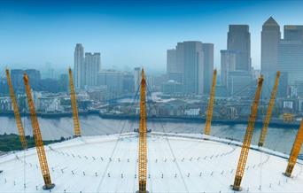 The white roof of The O2 with yellow pylons overlooking the River Thames and Canary Wharf