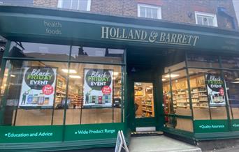 Outside Holland & Barrett in Woolwich. Showing a green shop-front with glass windows and posters.
