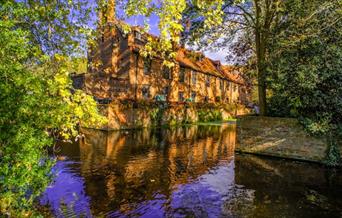 Looking over the moat at a colourful and sunny image of the Tudor Barn at Well Hall Pleasaunce Park and gardens