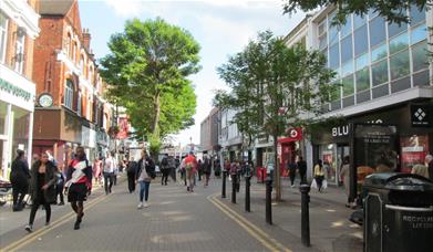Woolwich high street, a road with shops lined up on each side. Also showing benches and bins for public use.