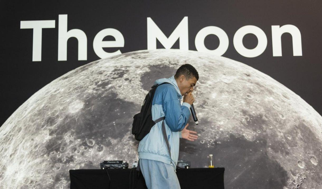 Youth artist with a mic in his hand facing right side in front of 'The Moon' banner.