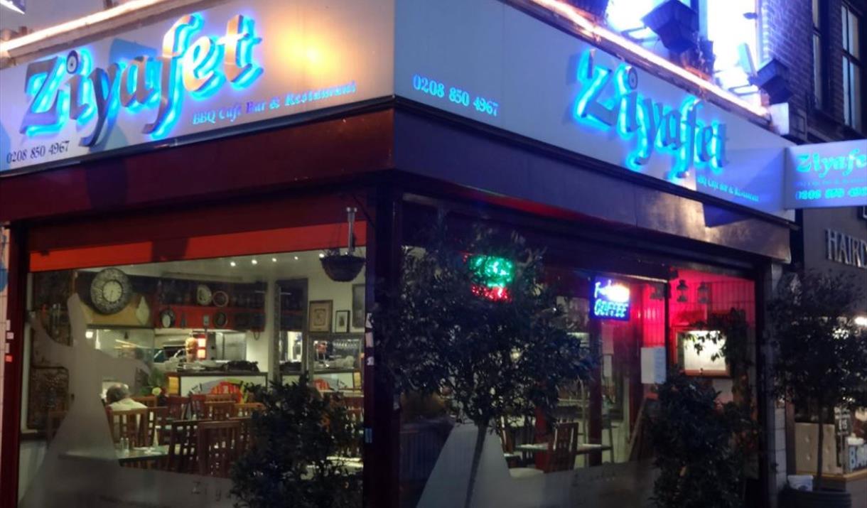 Night view of entrance of Ziyafet Restaurant exterior with big glass window around and name glowing with blue light in the background.
